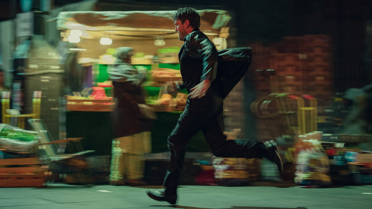 A man in a black jacket runs through a city street at night, with blurred lights and a market stall in the background, suggesting urgency in an urban environment.