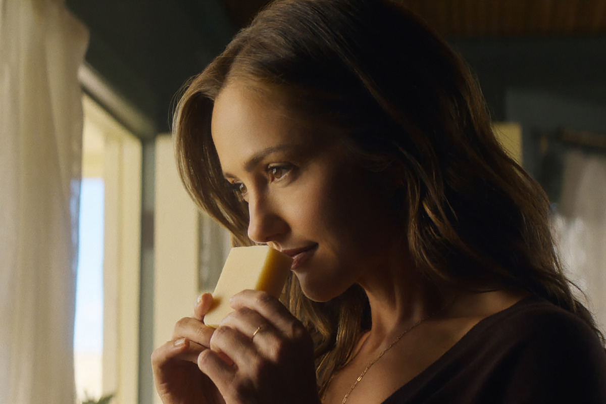 A woman stands indoors by a sunlit window, holding and smelling a piece of cheese, with soft natural light highlighting her face and a calm, cozy atmosphere surrounding her.