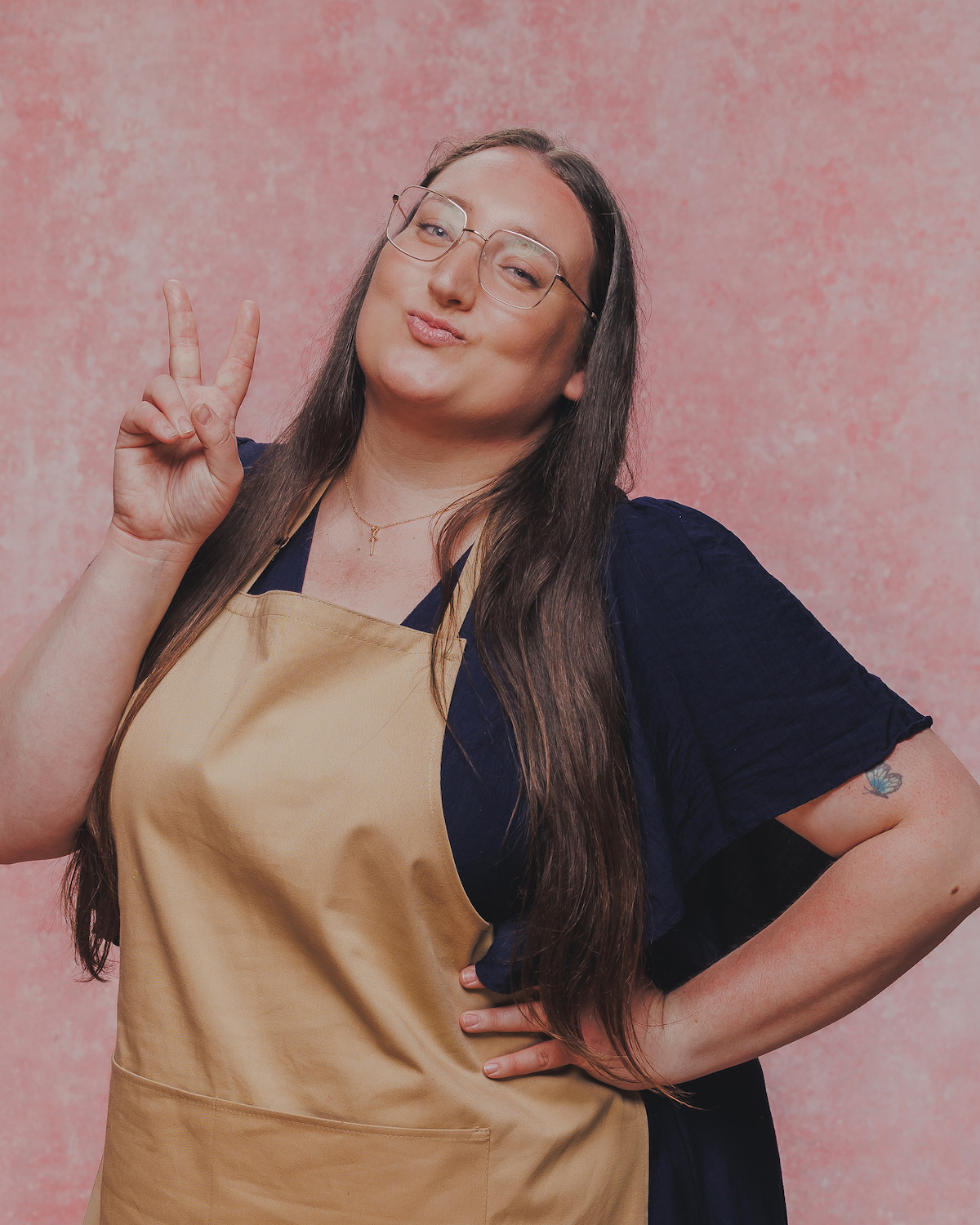 Person with long hair and glasses wearing a tan apron and navy dress poses with a peace sign and a playful expression in front of a pink textured background, standing confidently with one hand on their hip.