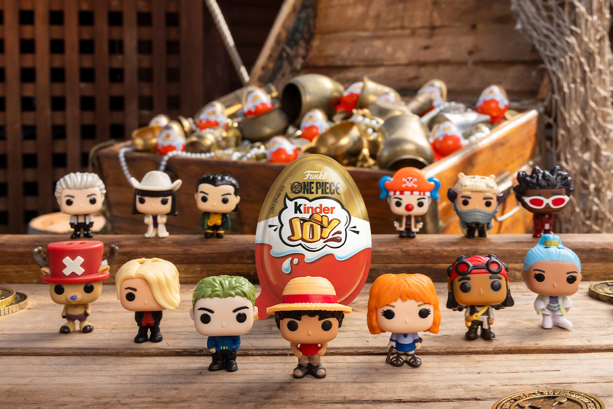 One Piece toy figures arranged on a wooden table with a Kinder Joy egg in the middle, pirate-themed chest in the background filled with gold eggs, all set in a rustic, wooden environment.