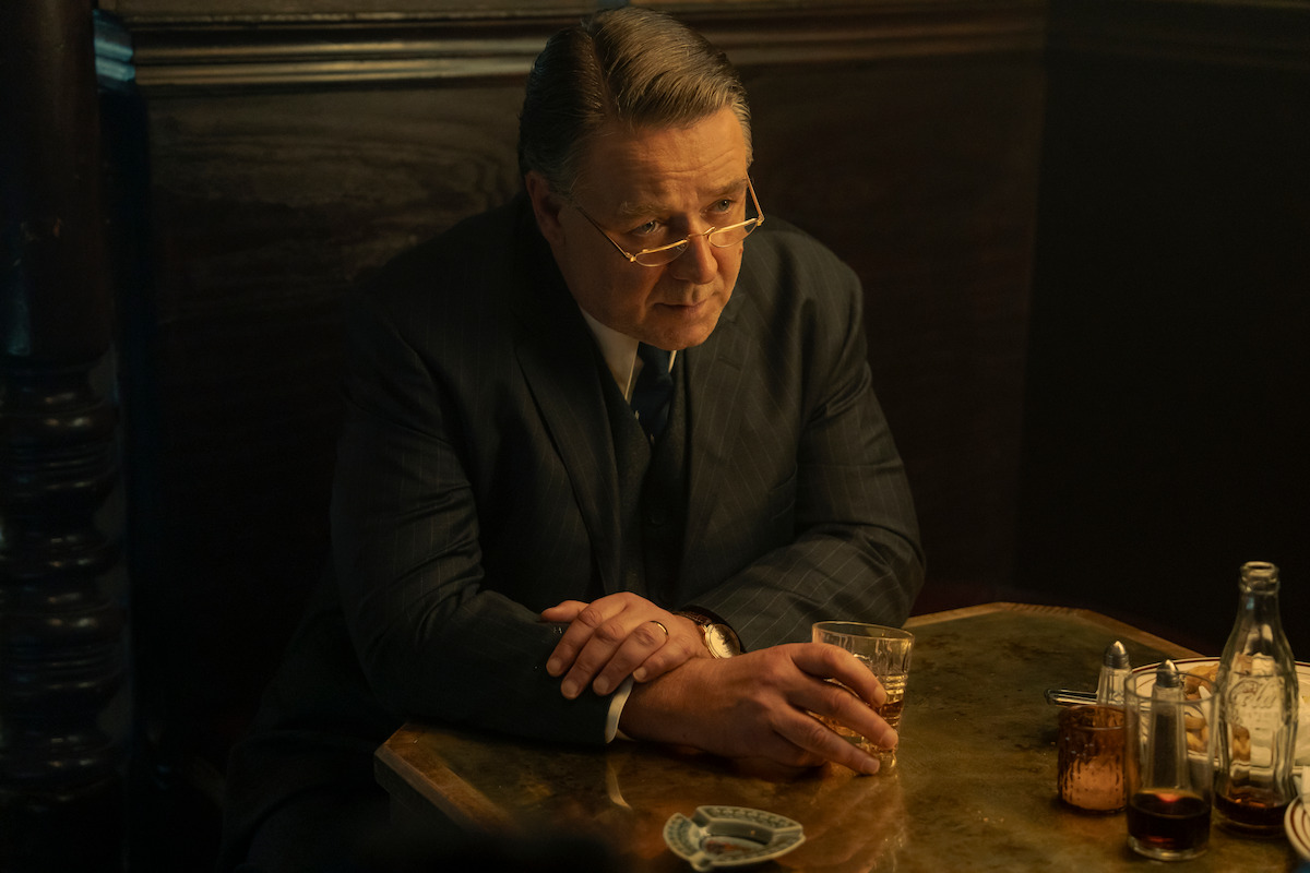 Man in a suit sits alone at a dimly lit bar or restaurant table, holding a glass of drink, with bottles and glassware on the table, appearing thoughtful or contemplative in a vintage setting.