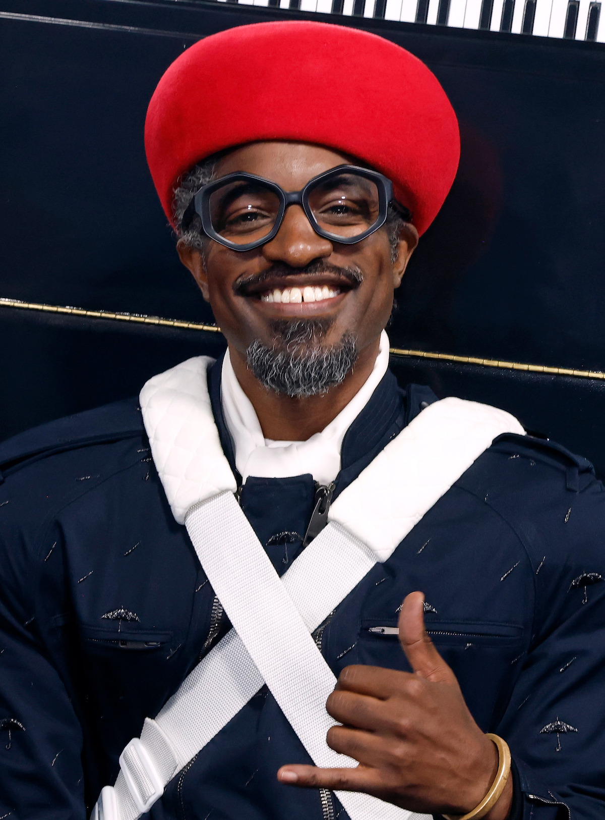 Smiling man wearing a red beret, large black glasses, and a dark jacket with white straps, posing with a hand gesture against a dark background.