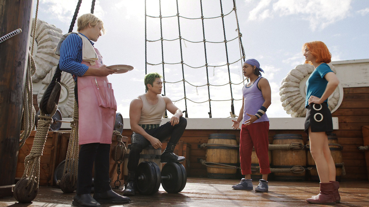 Four people in colorful outfits talking on the wooden deck of a ship, with coiled ropes, barrels, sails, and partly cloudy sky in the background during the day.