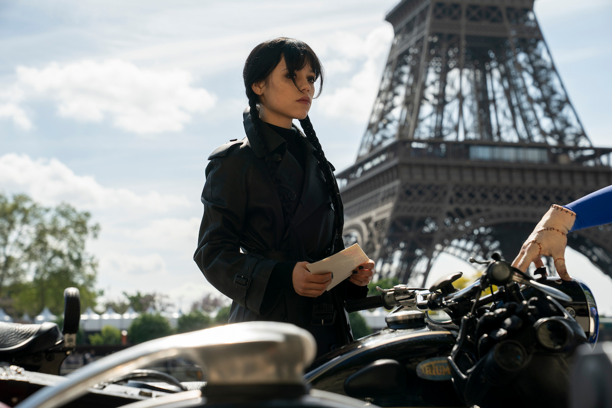 A person with dark braided hair stands next to motorcycles, holding papers, in front of the Eiffel Tower in Paris under a partly cloudy sky.
