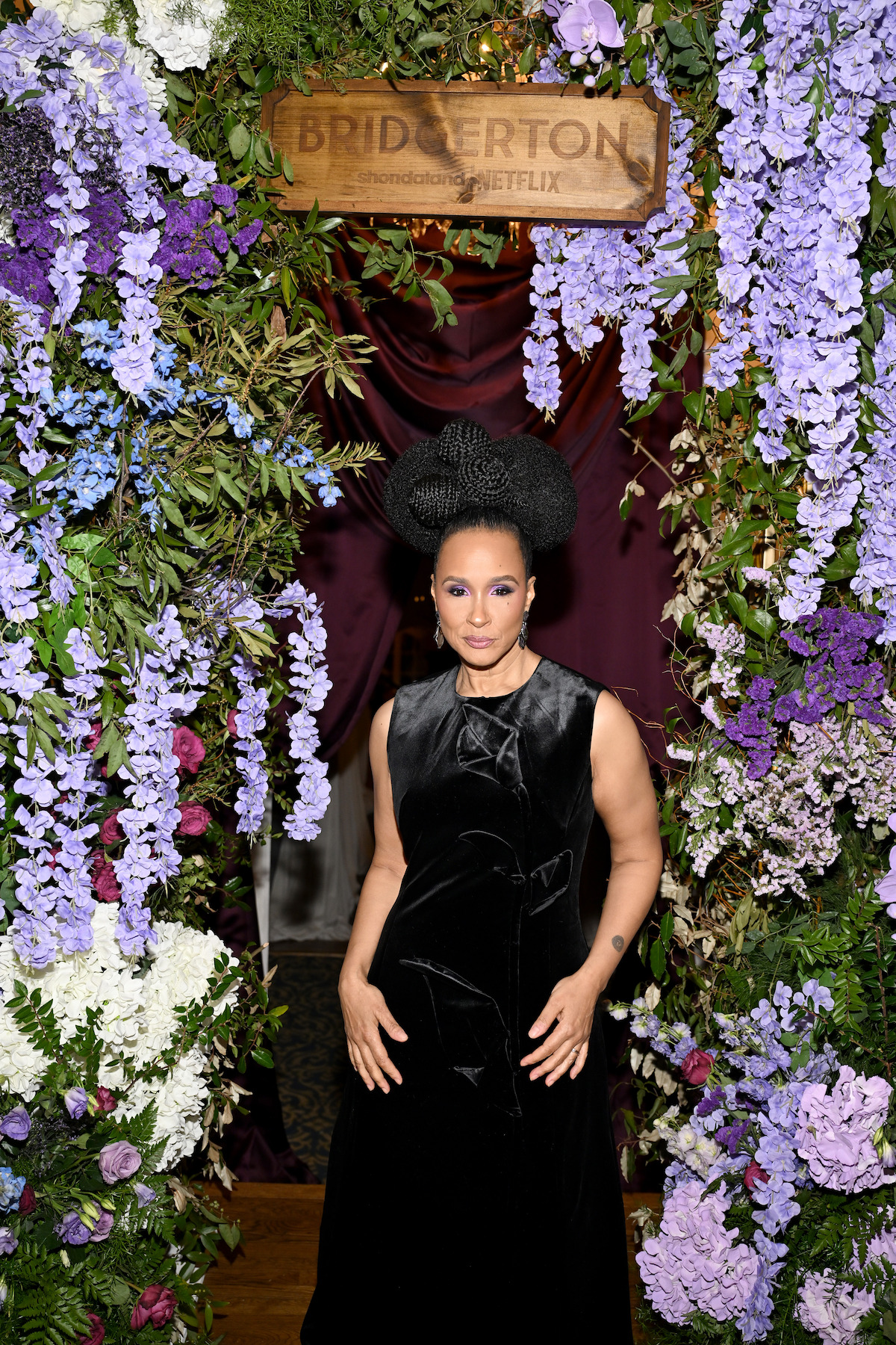 A woman in a black dress poses in front of a floral archway with purple and white flowers, under a Bridgerton and Netflix sign, at an elegant indoor event.