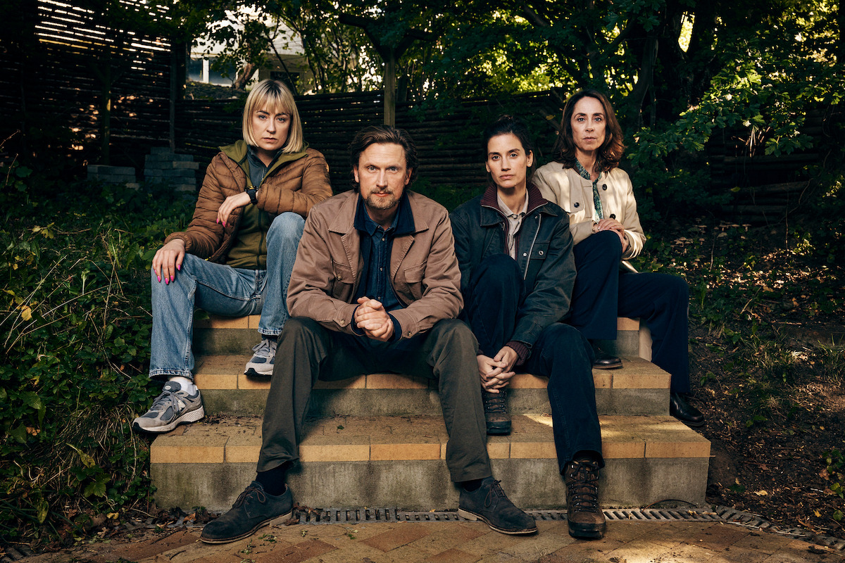 Four people sit on outdoor steps surrounded by greenery and wooden fencing, wearing casual jackets and pants, looking directly at the camera with serious expressions in a shaded garden setting.