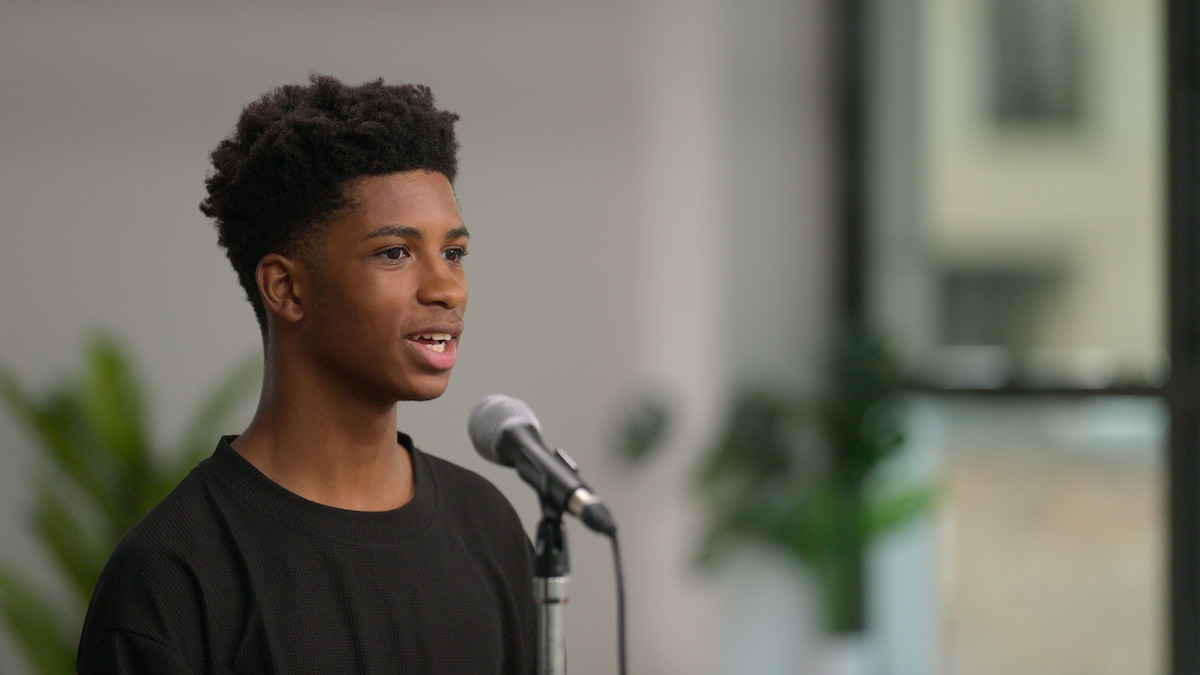 Teen boy speaking into a microphone indoors, with plants and a window in the background; bright, neutral setting, focused and confident mood.