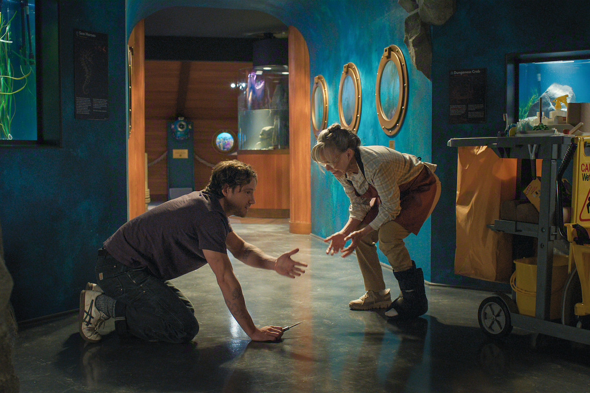 A man and a woman interact on the floor of a brightly lit aquarium hallway, surrounded by blue walls, exhibit windows, and cleaning supplies cart.