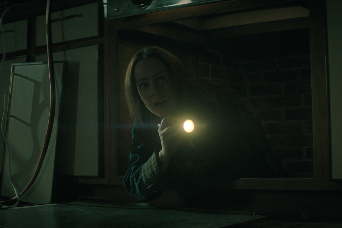 Jenna Malone as Claire, holding a flashlight, crawls through a dark crawl space or tunnel inside a dimly lit room, surrounded by wood and brick, creating a tense and mysterious atmosphere.