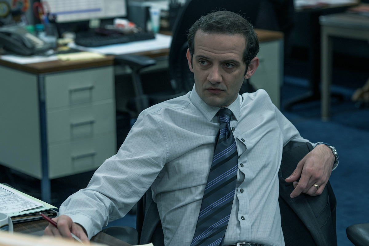 Man in dress shirt and tie sitting in an office environment at a desk, looking to the side with a serious expression. The office has folders, papers, and computer monitors on desks in the background.