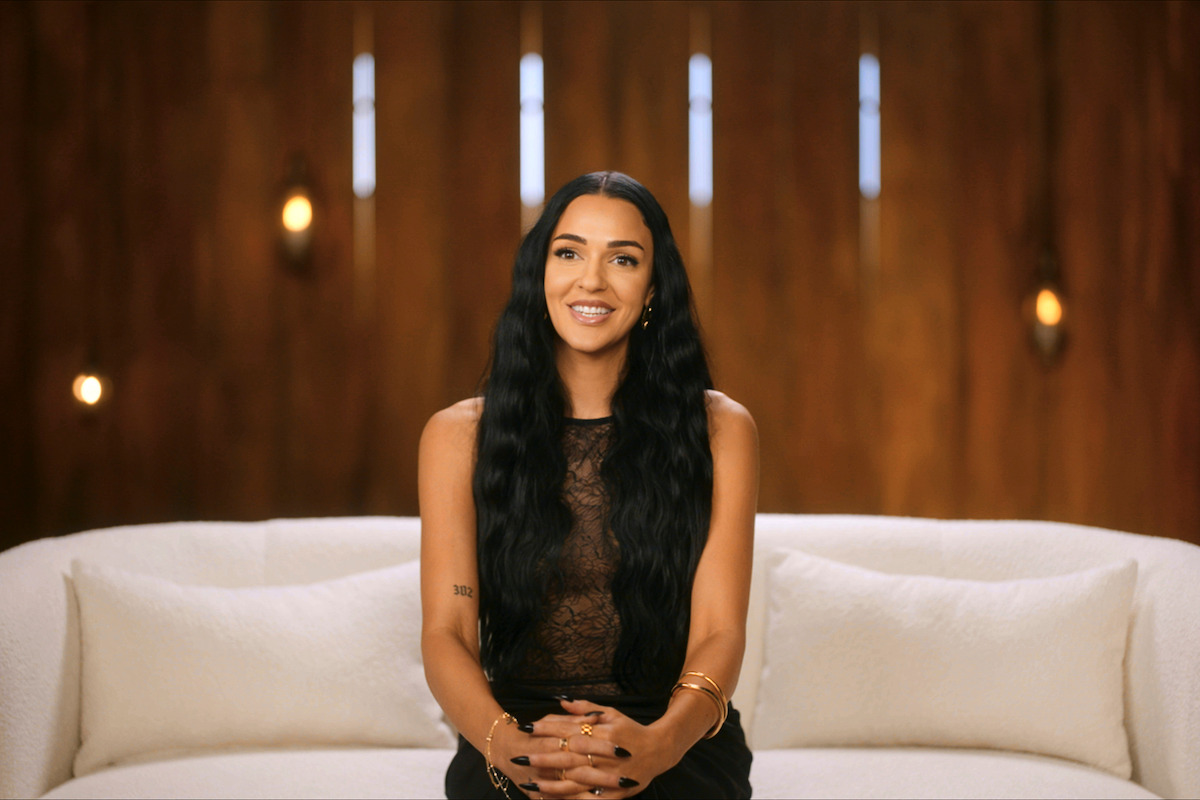 Woman with long dark hair sitting on a white couch, smiling, in a warmly lit room with a wooden wall and decorative lights in the background.