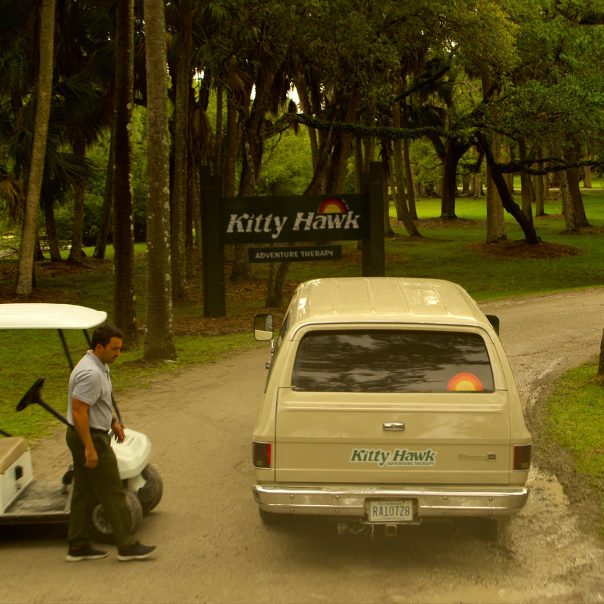 Homem está em pé ao lado de um carrinho de golfe perto de um carro bege em uma estrada cercada de árvores, com uma placa onde se lê “Kitty Hawk Adventure Therapy” visível no ambiente verde e exuberante do parque.