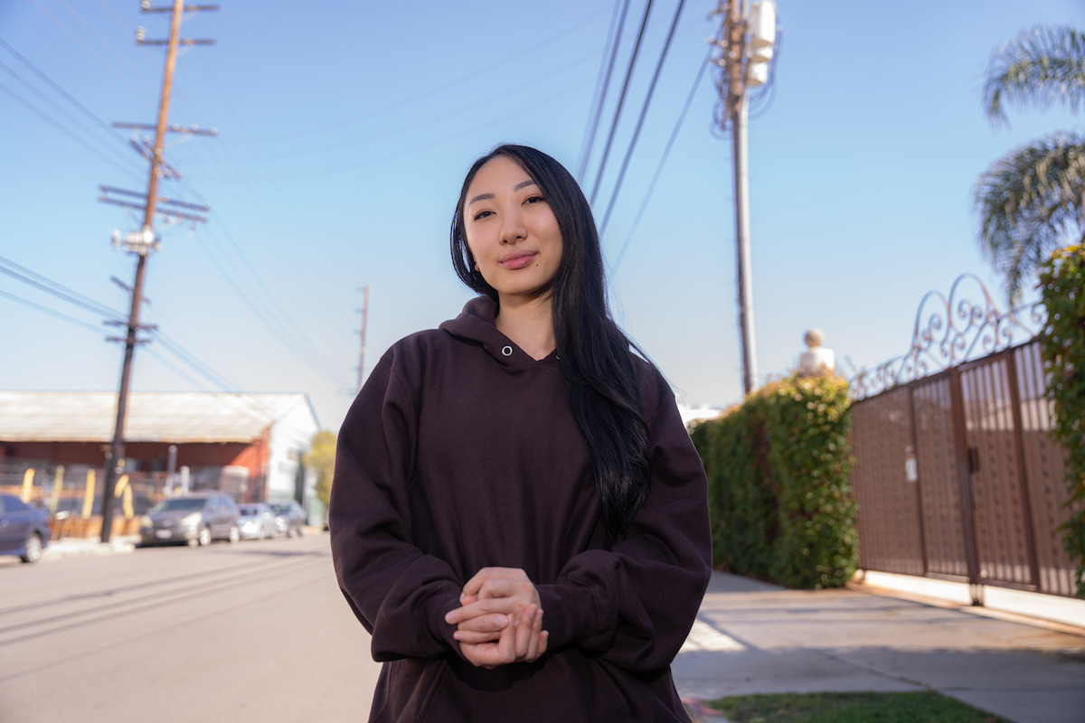 Woman in a brown hoodie standing on a sunny residential street with power lines, parked cars, palms, and a gated fence in the background.