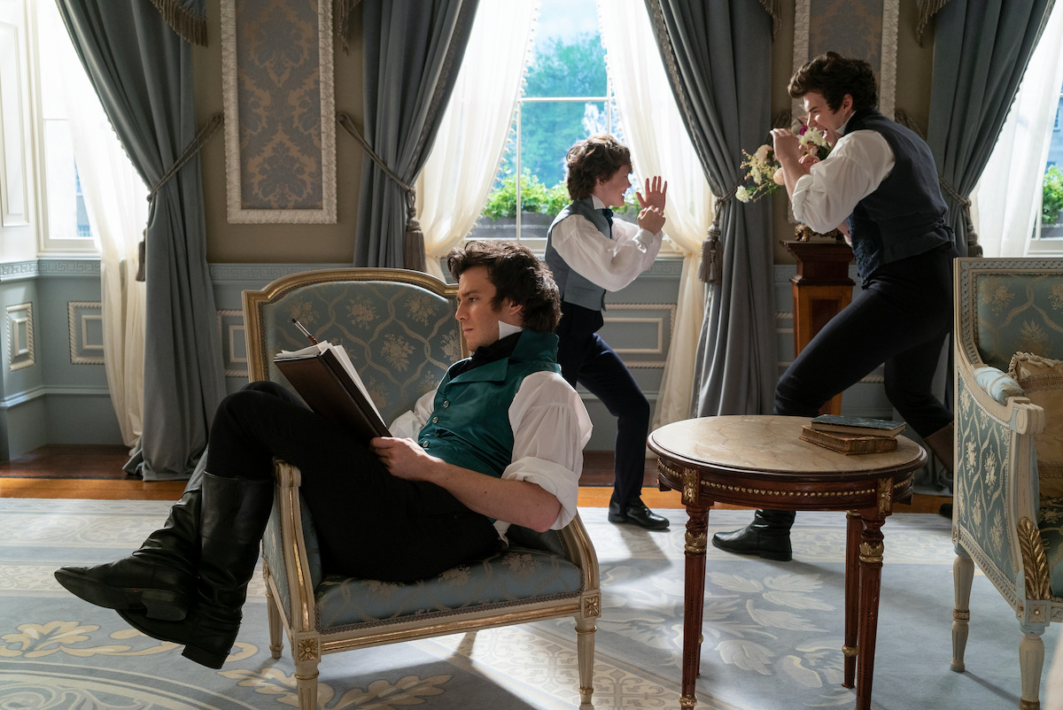 Three men in period clothing are in an elegant, ornate room; one sits reading, while the others act playfully near tall windows with heavy drapes and a view of greenery outside.
