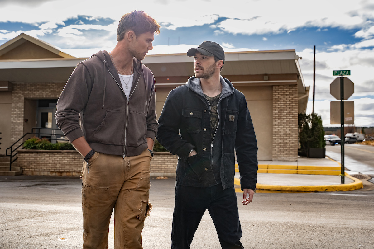 Two men walking and talking on a street in front of a building under a cloudy sky, dressed in casual outdoor clothing, with one in a brown hoodie and tan pants and the other in a black jacket and cap, urban daytime setting.