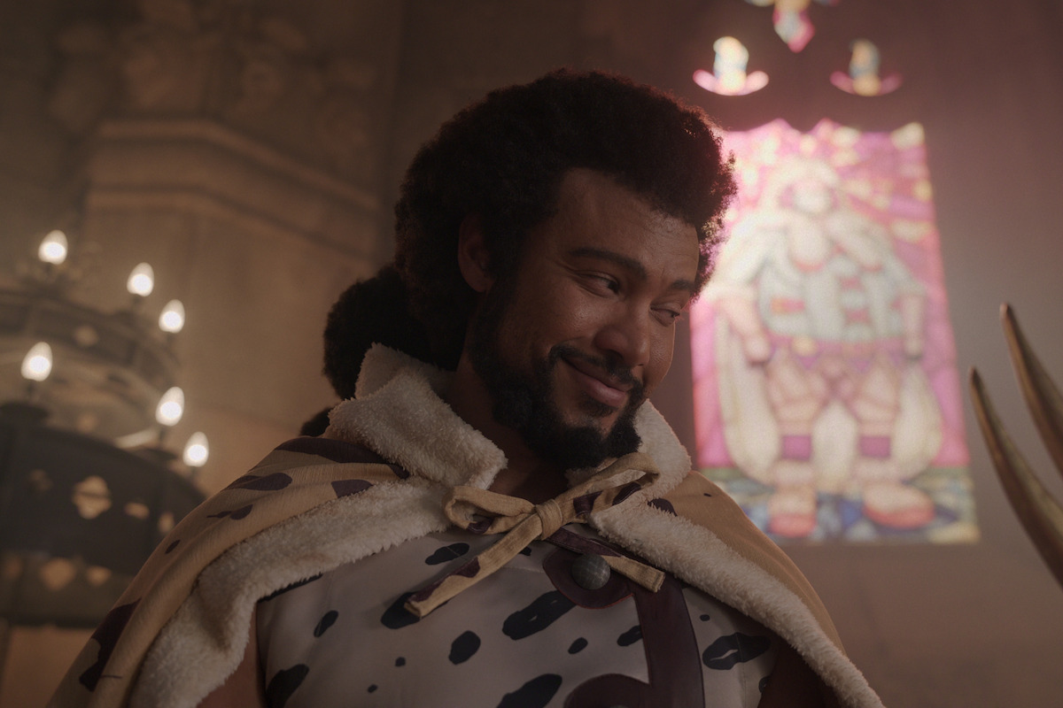 A man wearing a fur cape with a spotted pattern stands indoors, smiling slightly, with a colorful stained-glass window and soft, warm lighting in the background, evoking a medieval or fantasy setting.