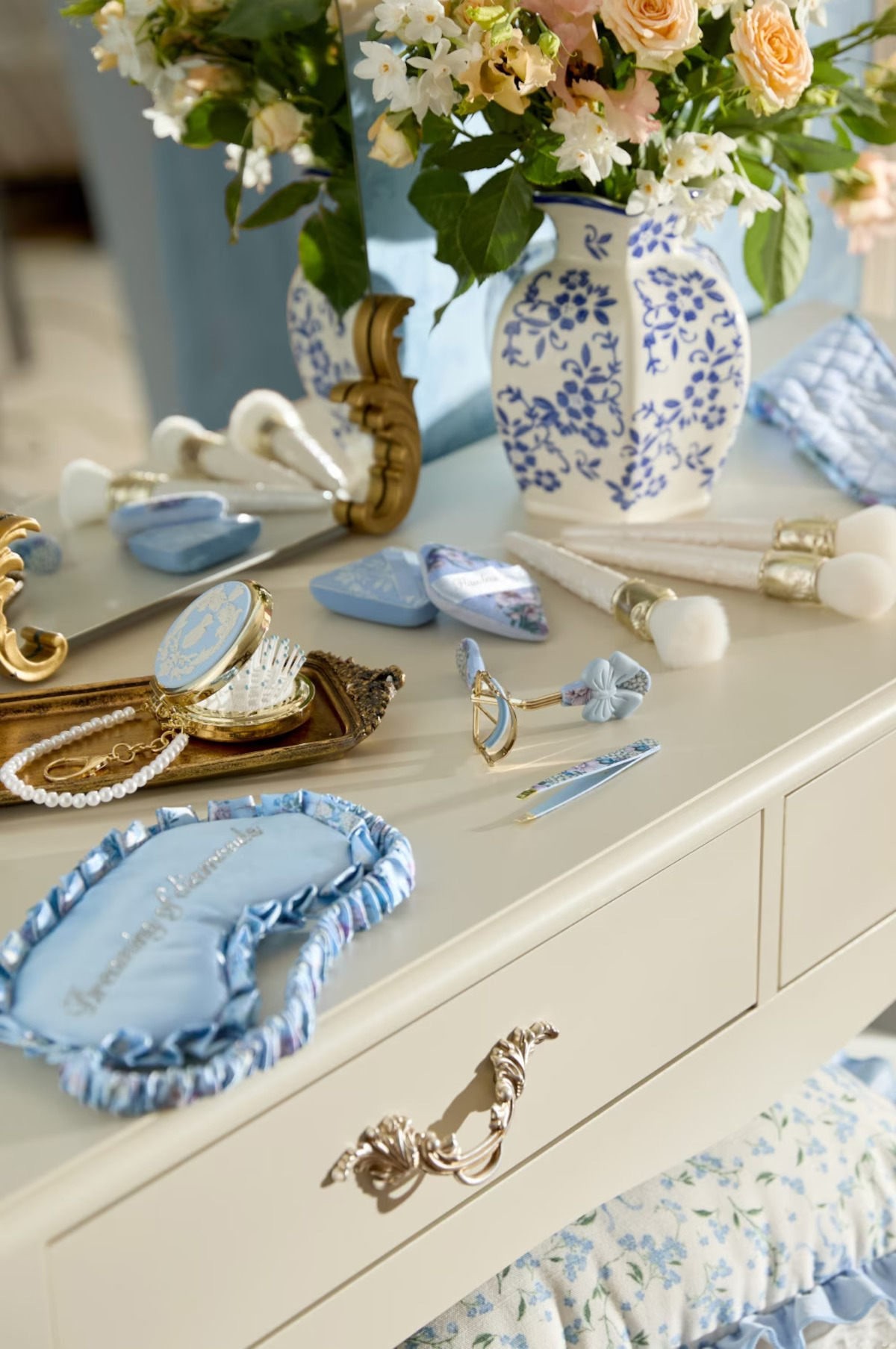 Vanity table with blue and white accessories, gold mirror, brushes, jewelry, sleep mask, and a floral vase, set in a bright, elegant bedroom with a feminine, vintage feel.