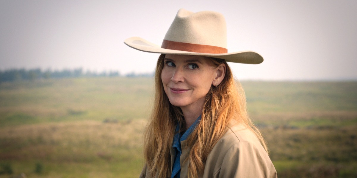 A woman with long hair wearing a tan cowboy hat and tan coat stands outdoors in a grassy open field, with a faint tree line and hazy sky in the background.
