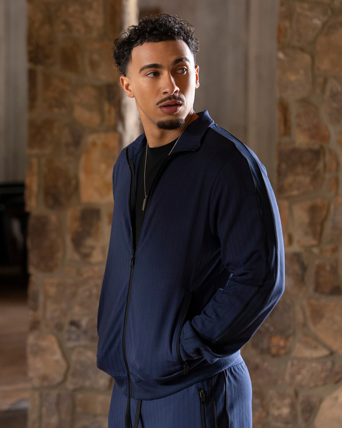 Man in a navy tracksuit standing indoors near a stone wall, looking to the side with hands in pockets, natural light filtering in, relaxed atmosphere.