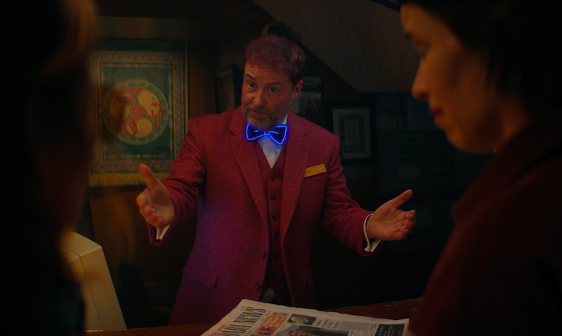 A man in a red suit with a glowing blue bow tie gestures behind a counter, speaking to two people holding a newspaper in a warmly lit, eclectic room, creating a mysterious and engaging atmosphere.