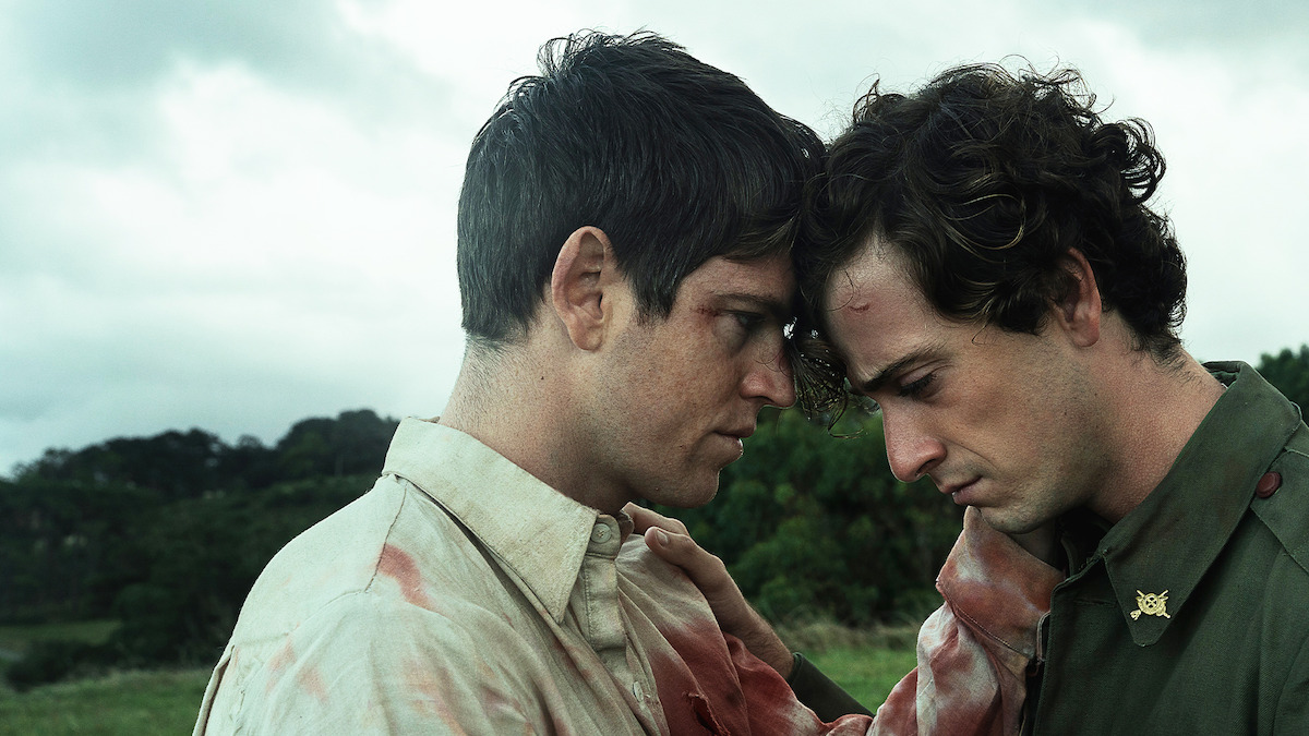 Two injured men with bloodied clothes stand close together in an emotional moment, foreheads touching, in an outdoor grassy field under a cloudy sky, suggesting a tense or dramatic scene.