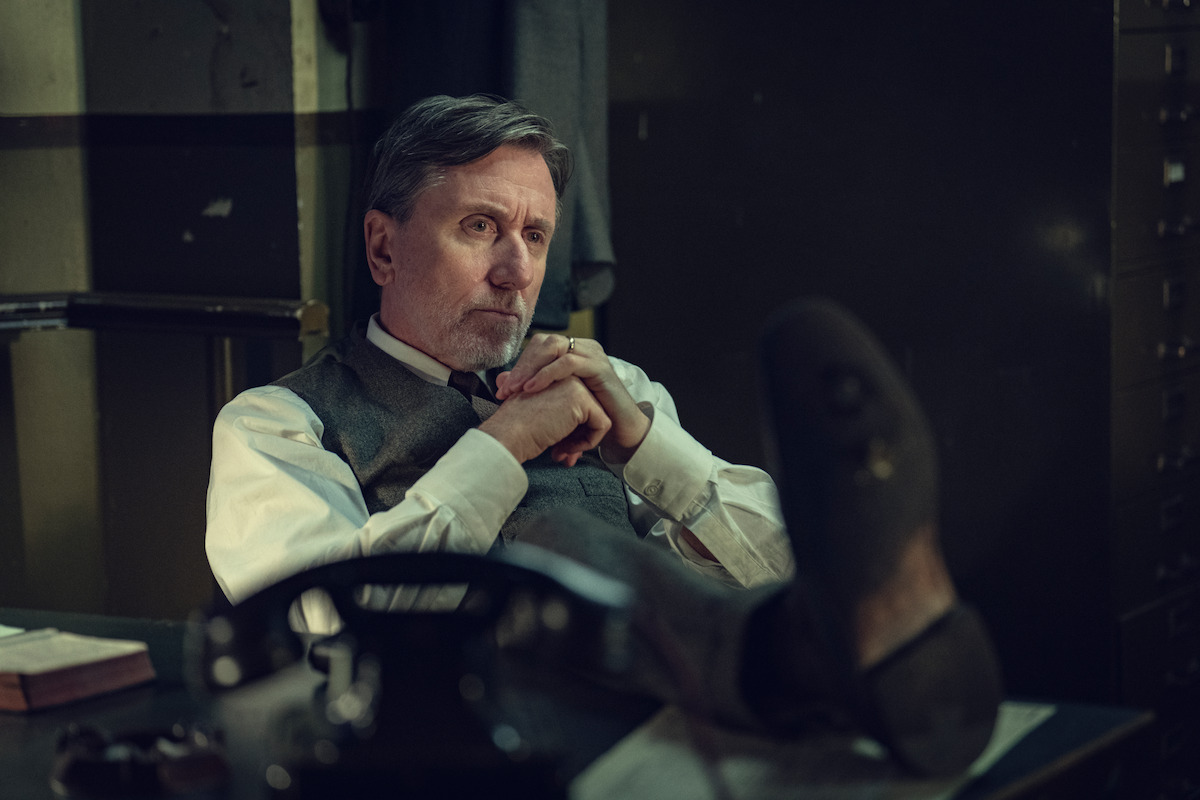 Man in dress shirt and vest sits at a desk in a dimly lit office, leaning back with feet up, hands clasped, looking thoughtful. Papers, a rotary phone, and a book are on the desk. The setting appears vintage or mid-20th century.