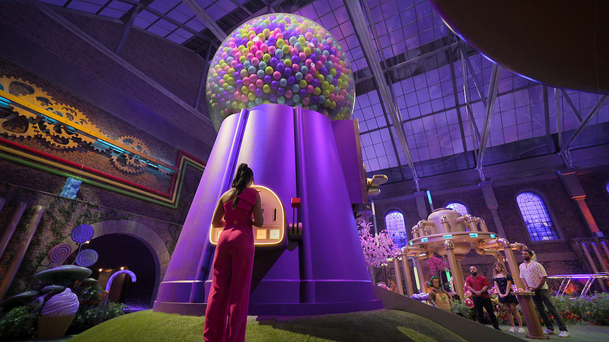 A woman in a pink outfit stands before a giant purple gumball machine under a glass ceiling in a whimsical, candy-themed indoor exhibit with other people and colorful decorations in the background.