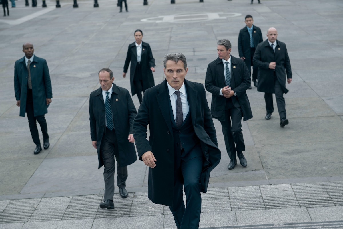Rufus Sewell as Hal Wyler walks up the steps of a government building with a group of formally dressed walking behind him.