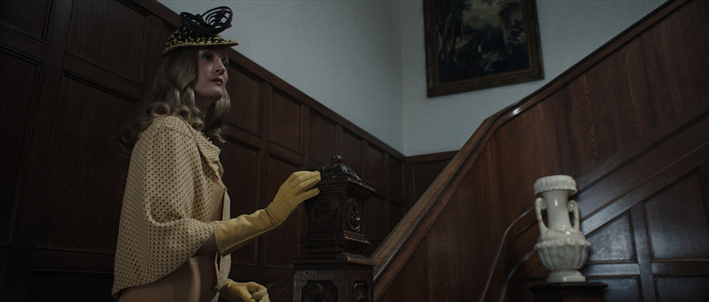 A woman dressed in vintage clothing, including a yellow hat, gloves, and cape, stands at the bottom of a wooden staircase in an elegant, wood-paneled interior.