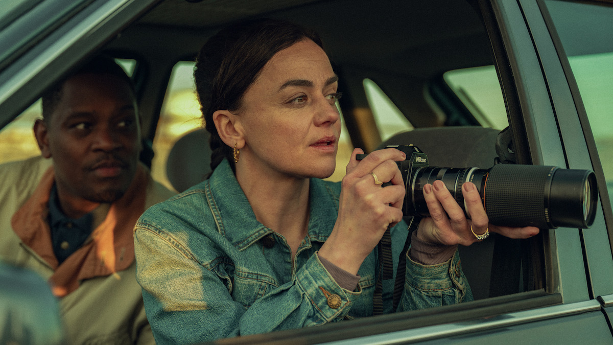 Two people sit in a car, with a woman in a denim jacket holding a camera with a large lens, looking intently outside as if observing or photographing something, while a man sits beside her, watching her actions.