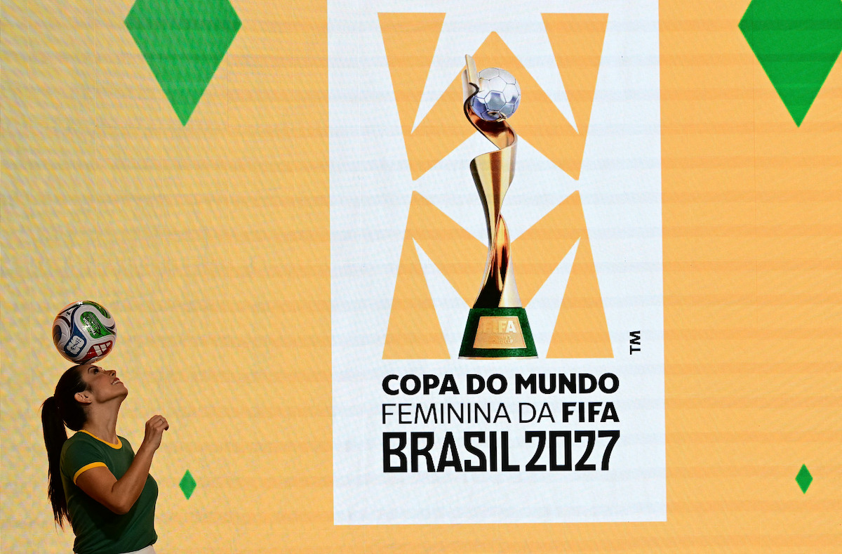 A woman balances a soccer ball on her head in front of a bright yellow wall displaying the FIFA Women’s World Cup Brazil 2027 logo and text, with green diamond shapes in the environment.