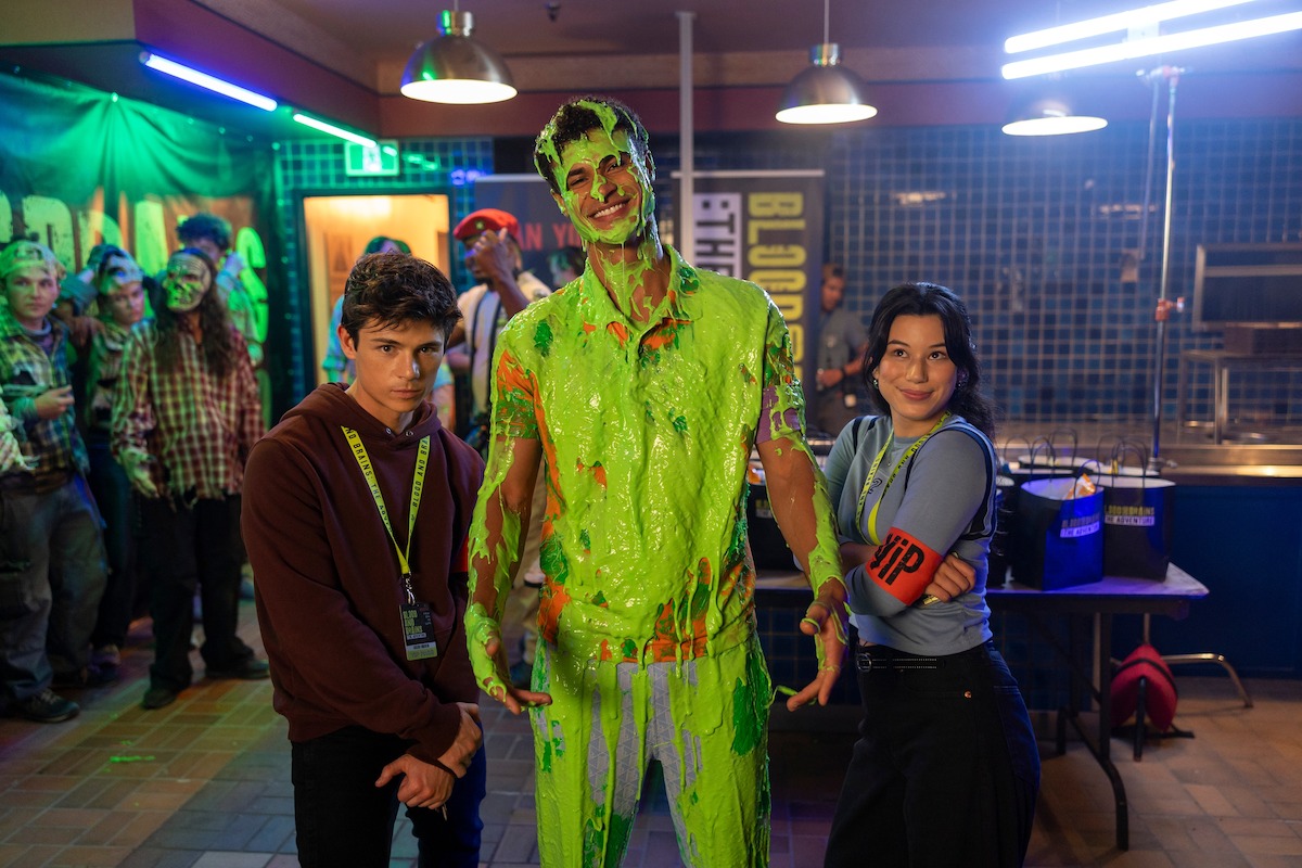 Three people at a lively indoor event; one is covered in green slime, while the others pose and smile. The background has neon lighting, event signage, and more people, some also slimed, suggesting a fun, themed gathering.