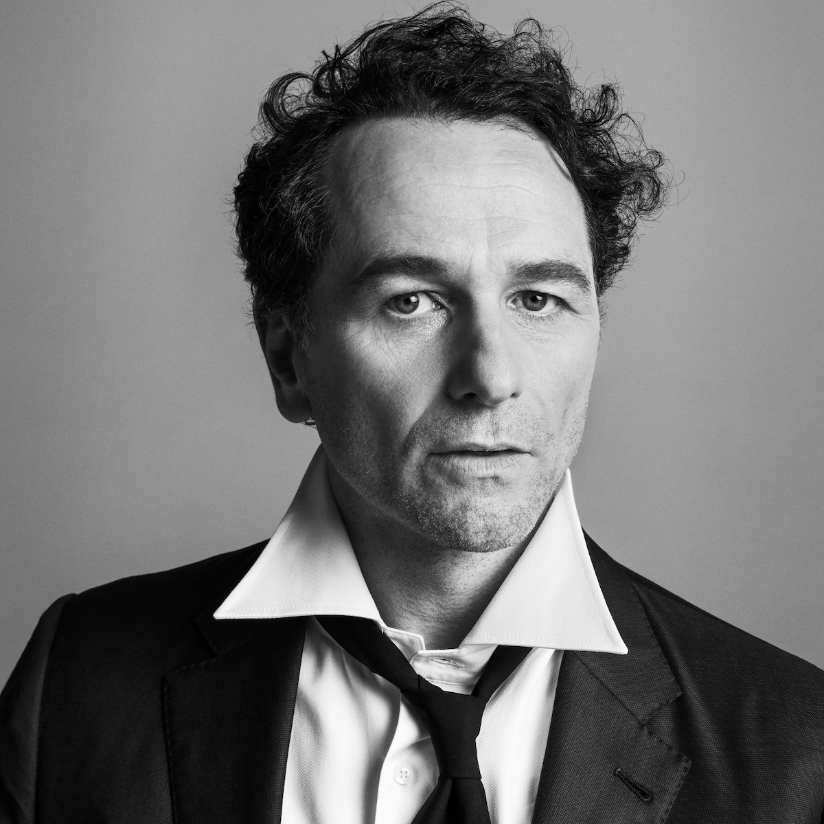 Black and white portrait of a man in a suit with an open collar and loosened tie, neutral expression, messy hair, studio background, moody and introspective atmosphere.