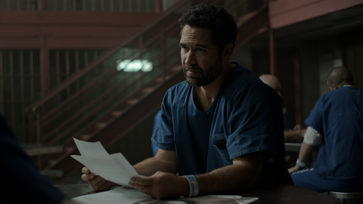 Man in blue prison uniform sits at a table holding papers in a dimly lit jail common area, with other inmates in the background and metal staircases visible.