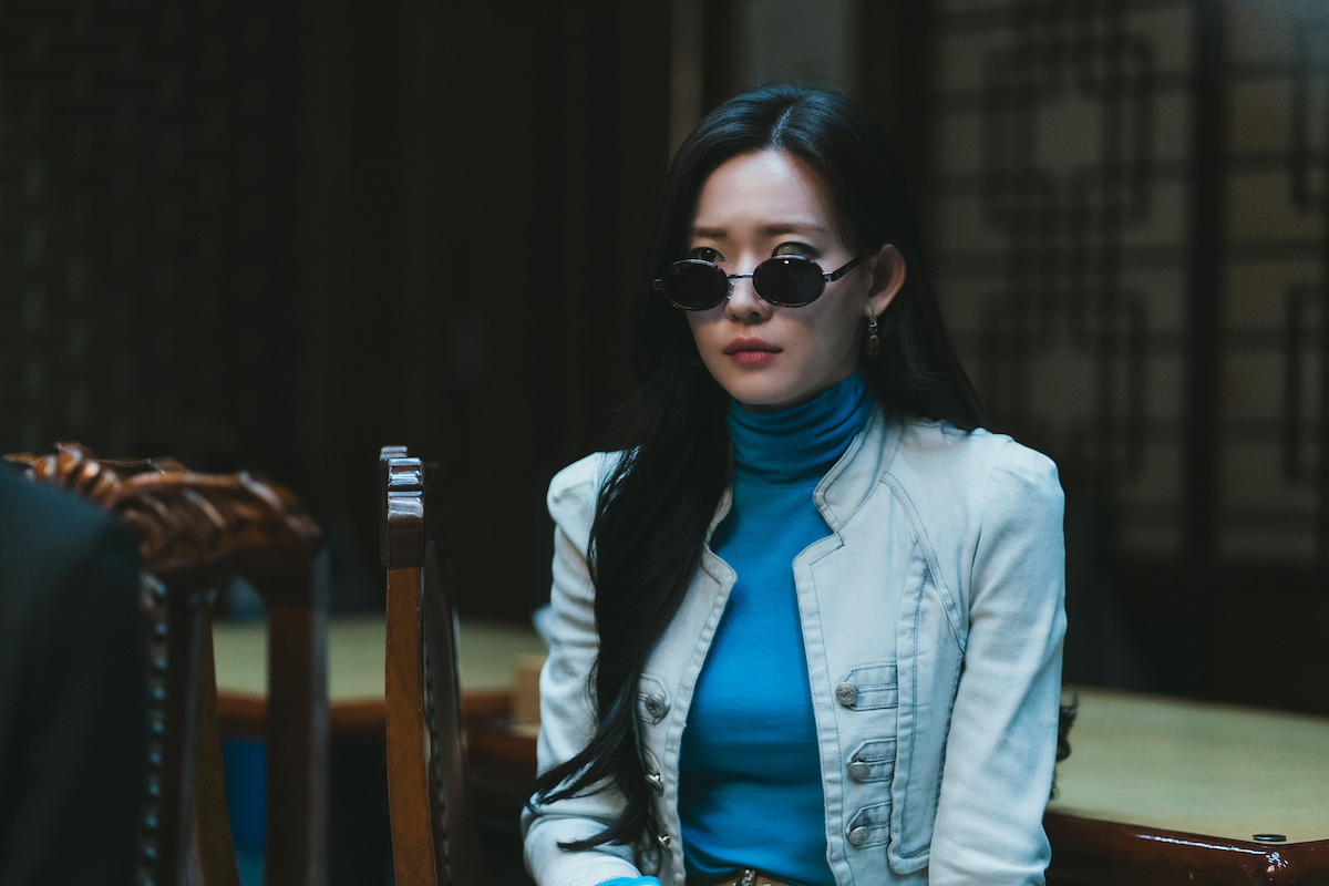 Woman with long dark hair wearing sunglasses, a blue turtleneck, and a white blazer sits indoors at a table with wooden chairs in a dimly lit room with traditional patterned wooden screens in the background.