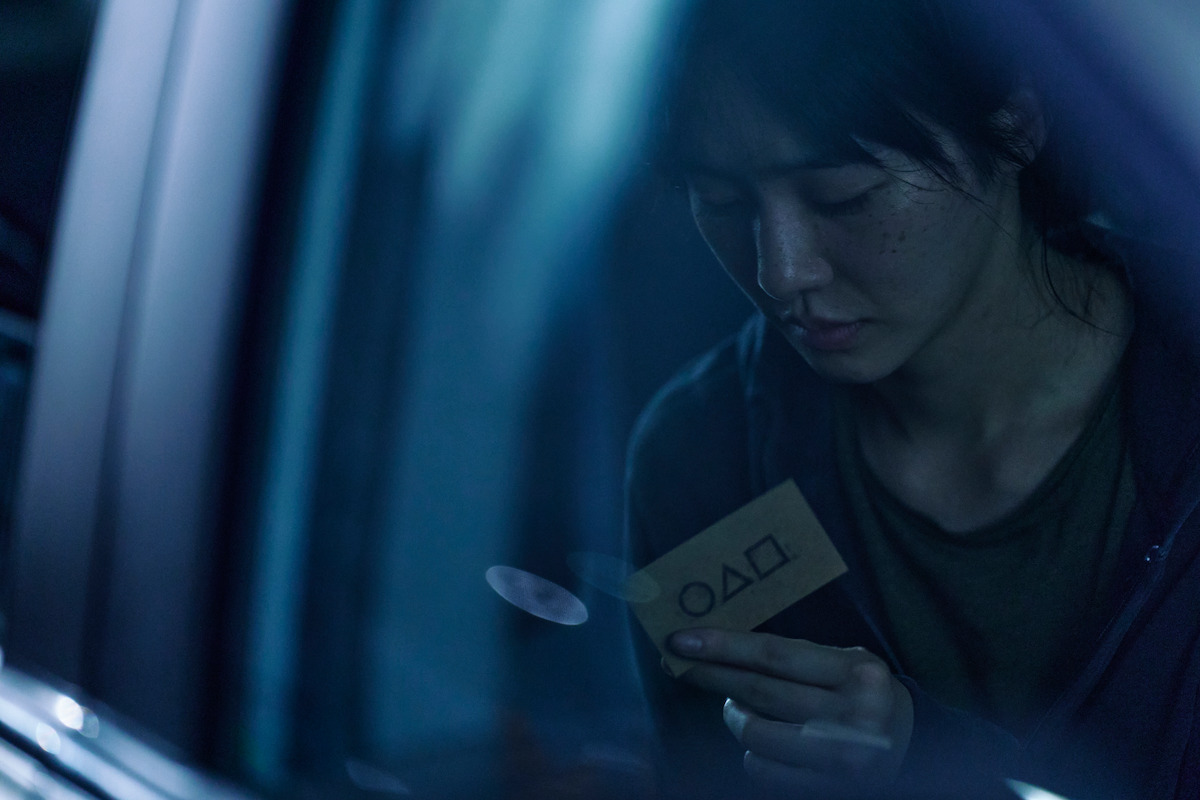 Reflection in a car window of a woman looking at a small card with a circle, triangle, and square on it in season 2 of ‘Squid Game’