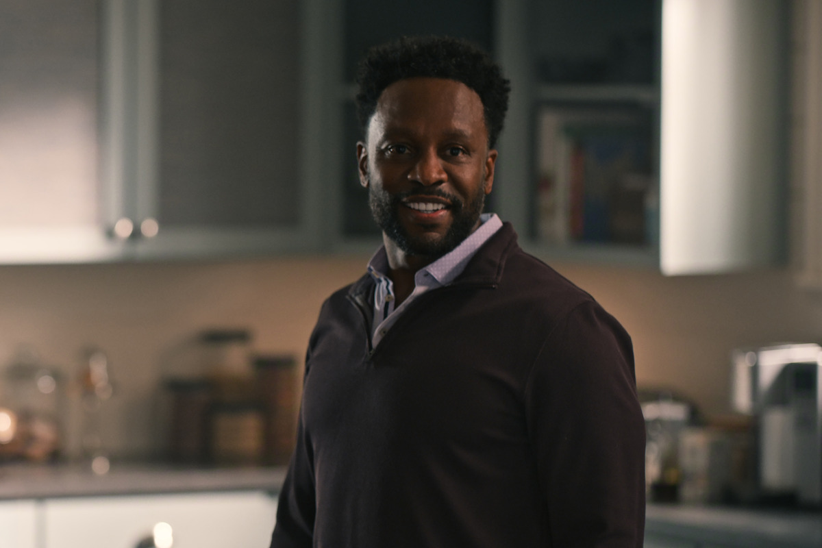 Smiling man with a trimmed beard standing in a modern kitchen, wearing a dark pullover over a collared shirt, with shelves, cabinets, and kitchen appliances in the background.