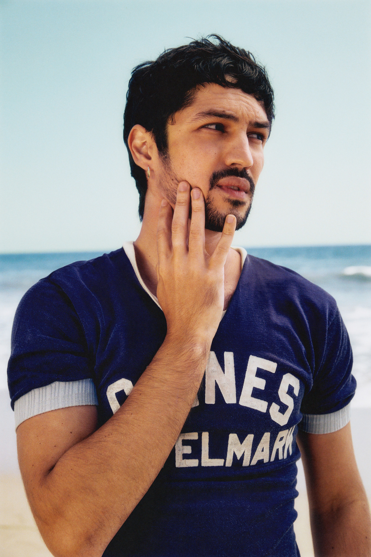 Gabriel Leone wears a navy shirt with a white collar and sleeves, and pulls at his cheek.