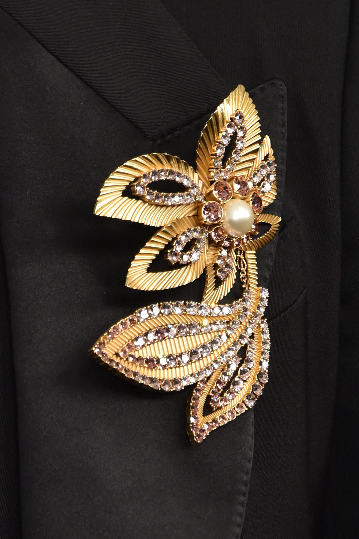 Gold and jeweled floral brooch with a pearl center pinned to a black suit lapel, showcasing intricate leaf designs, sparkling gemstones, and elegant metallic detail in a formal setting.