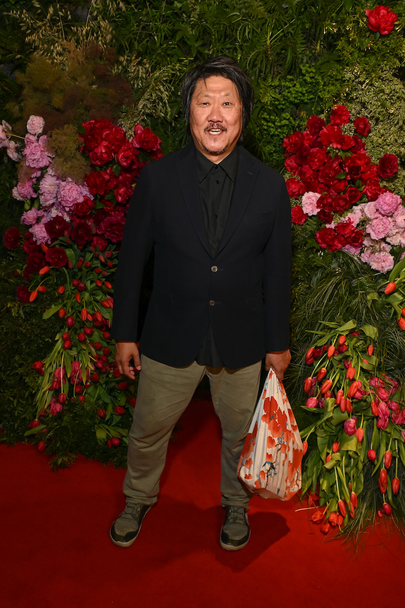 Man in a dark blazer and olive pants stands on a red carpet, holding an orange and white bag, with a lush floral backdrop featuring red, pink, and purple flowers and greenery. Indoor event setting.