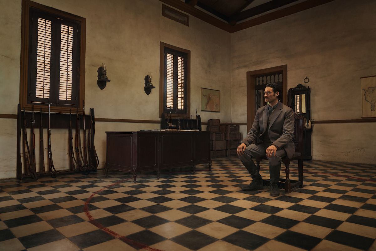 A man in a suit sits on a chair in a sparsely furnished, old-fashioned room with checkered floor, rifles against the wall, tall windows with shutters, wooden furniture, and muted lighting creating a somber atmosphere.