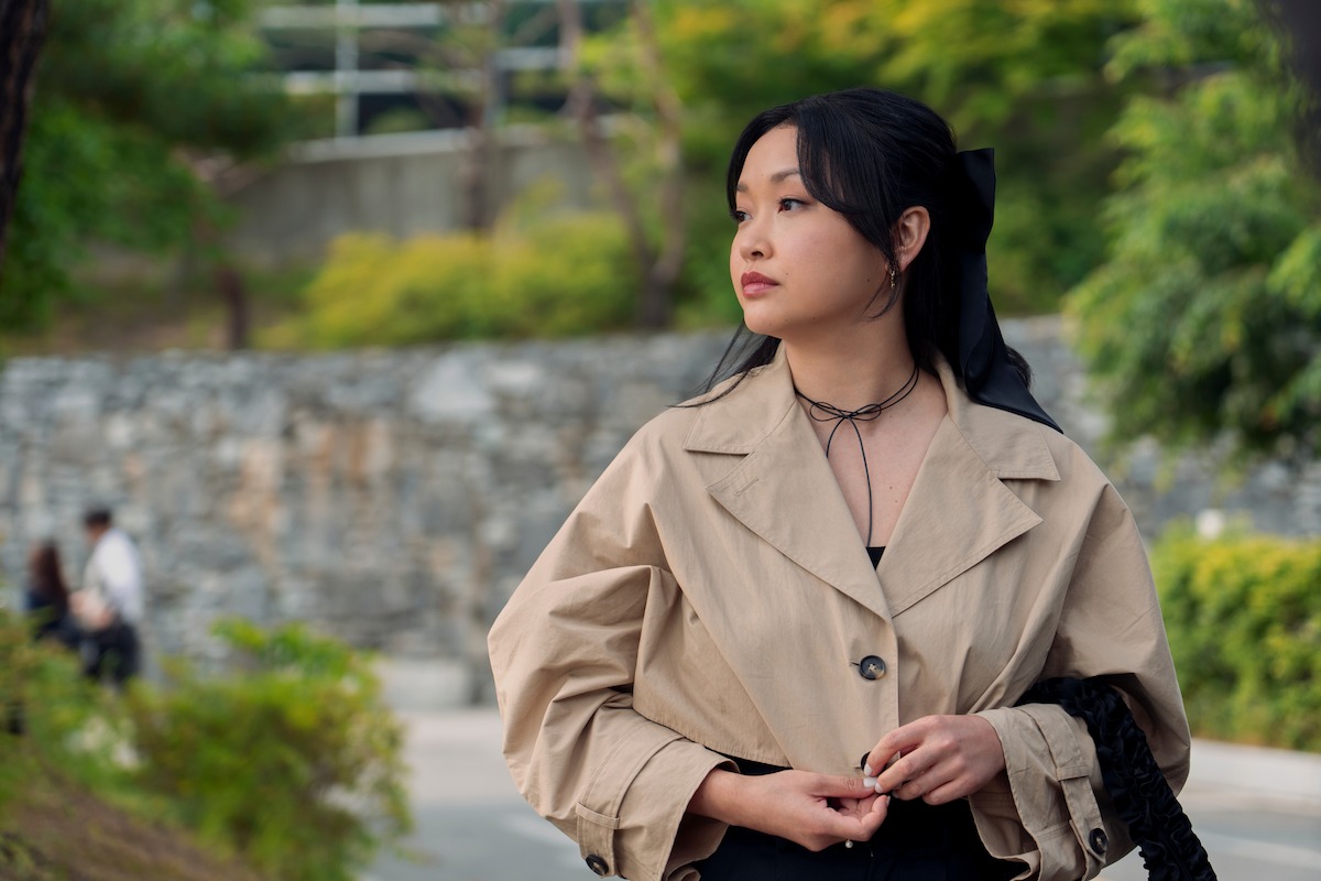 Woman in beige coat standing outdoors in a park-like setting with green trees, stone wall, and soft natural light, looking to the side thoughtfully.