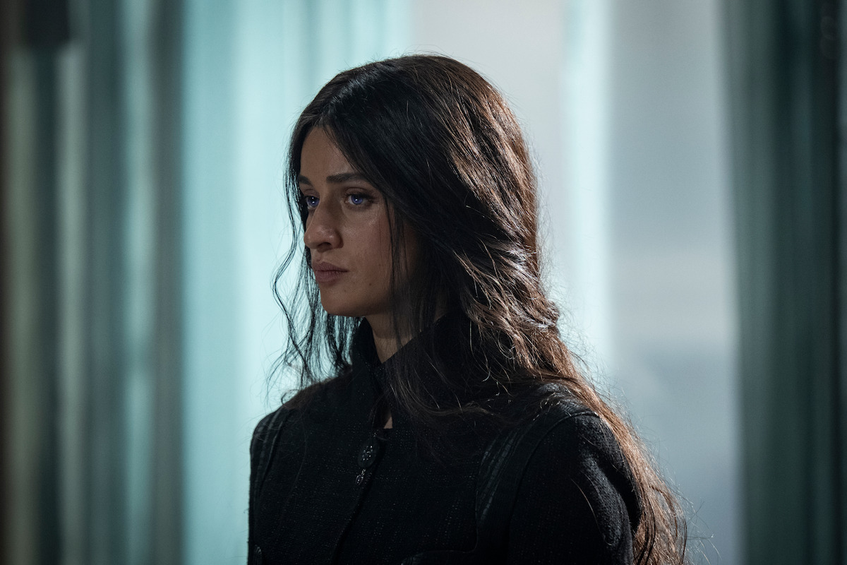A woman with long dark hair stands indoors in soft, diffused light, wearing a dark outfit. She appears serious and contemplative, with a somber mood reflected by the muted background and calm environment.