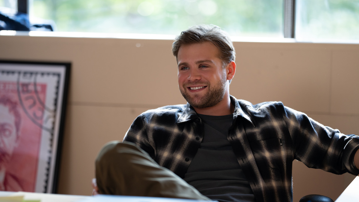 Smiling man in plaid shirt sits casually in a sunlit office, with a framed piece of artwork and window in the background.