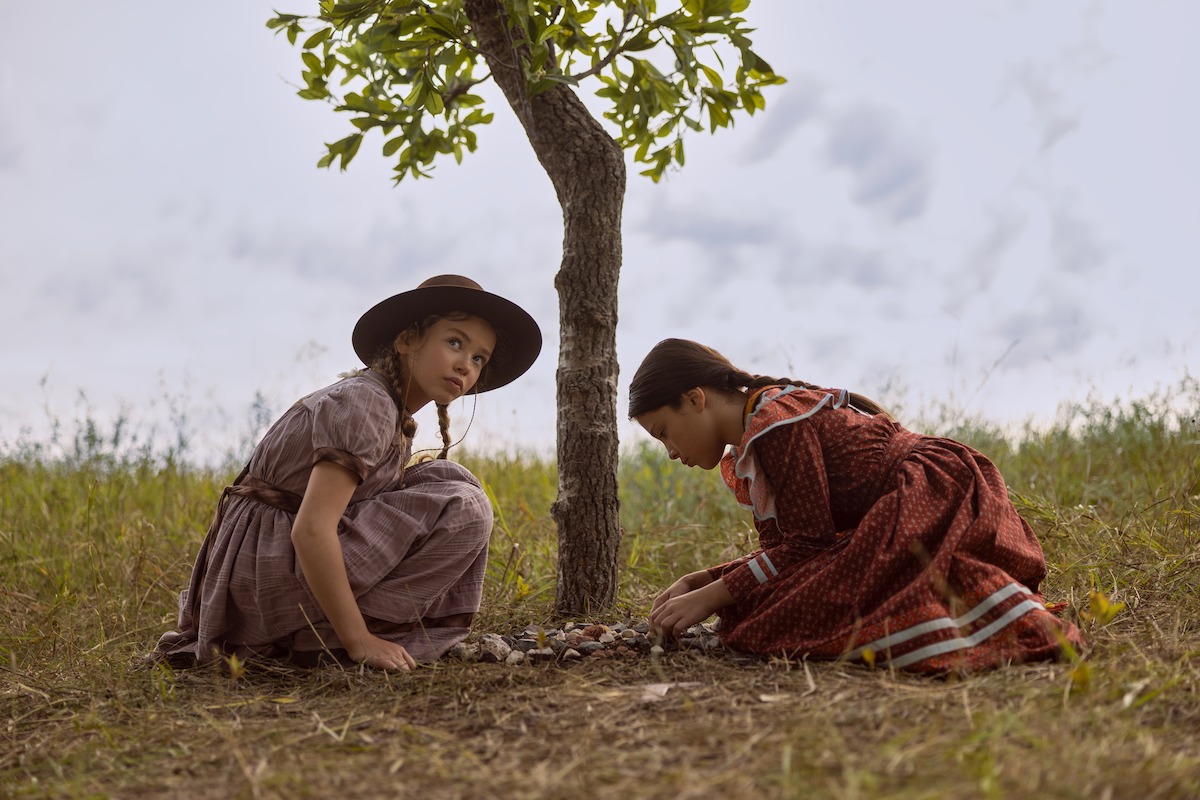 Two girls in vintage dresses crouch by a young tree in a grassy field, arranging stones at its base under a cloudy sky, evoking a peaceful, historical outdoor scene.