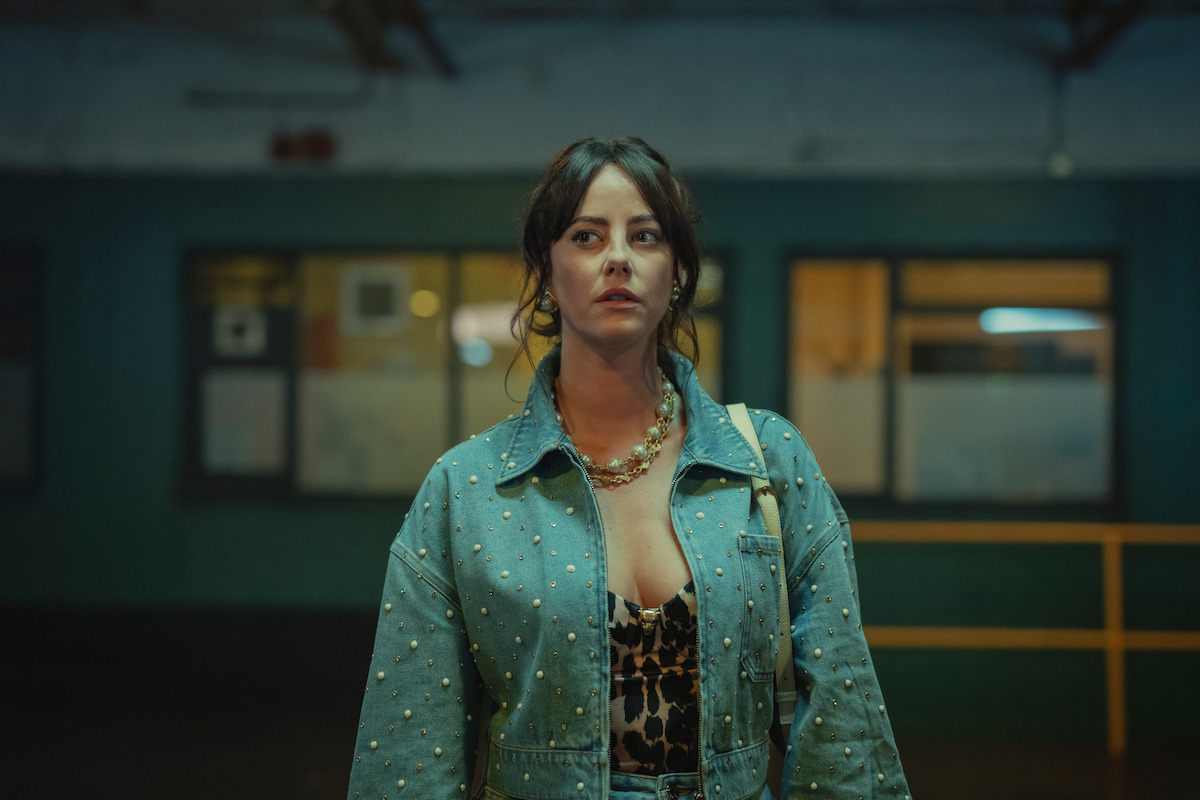 Woman with dark hair in a pearl-studded denim jacket and leopard print top standing indoors at night in a dimly lit industrial or urban setting with blurred windows and a yellow railing in the background.