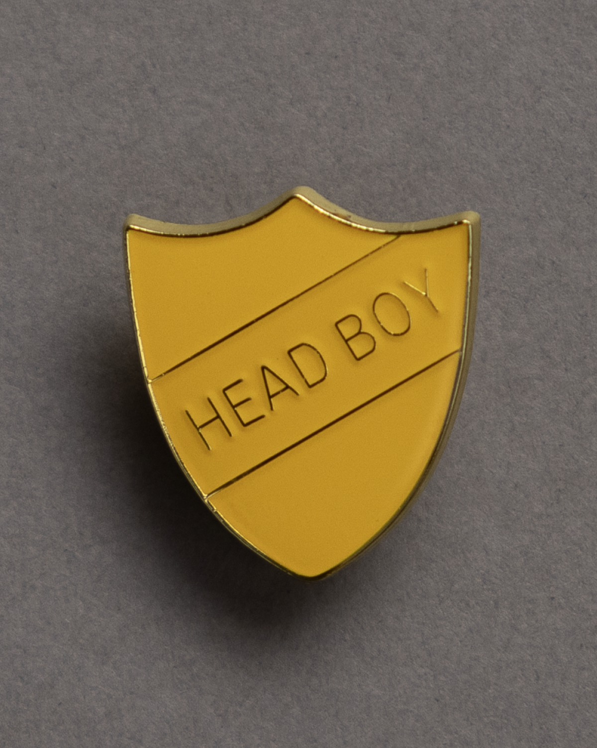 Yellow shield-shaped enamel pin with the words "HEAD BOY" embossed on it, positioned against a plain gray background.