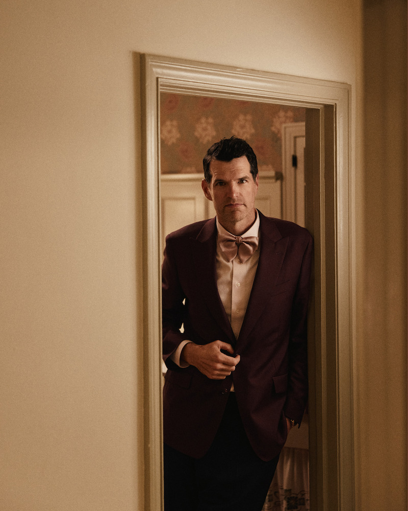 Man in a burgundy suit jacket and bow tie stands in a doorway of a warmly lit, vintage-style interior with patterned wallpaper in the background.