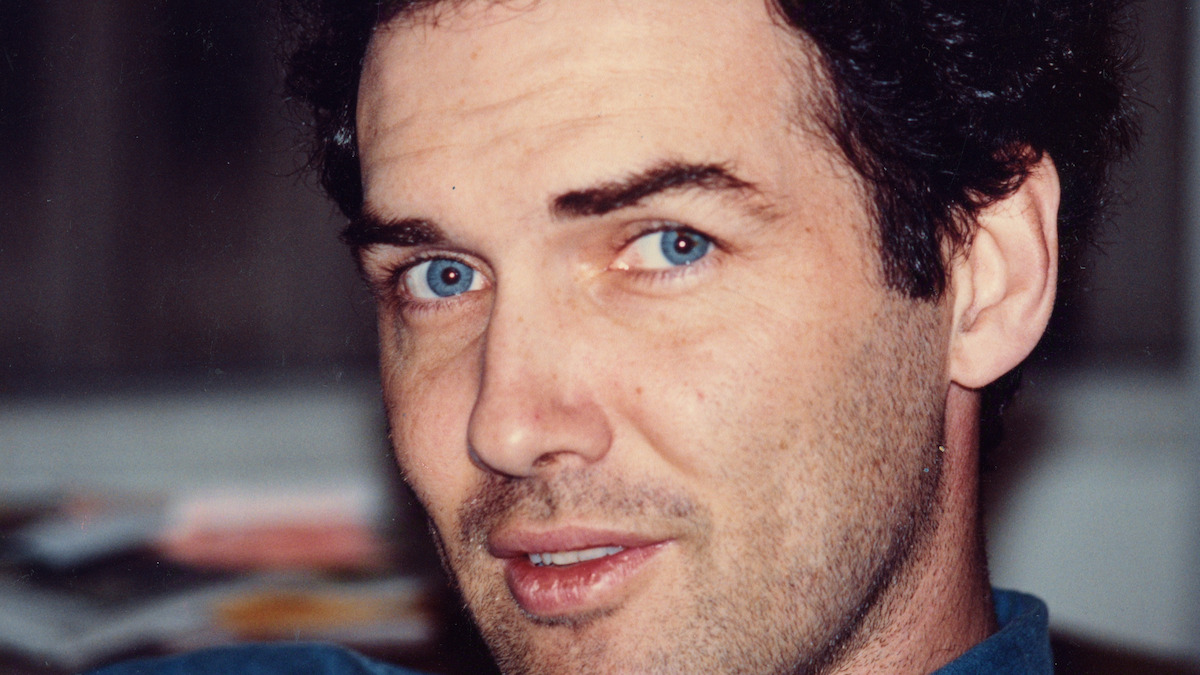 Man with curly dark hair and blue eyes looks at the camera indoors, with blurred background suggesting a casual home or office setting.
