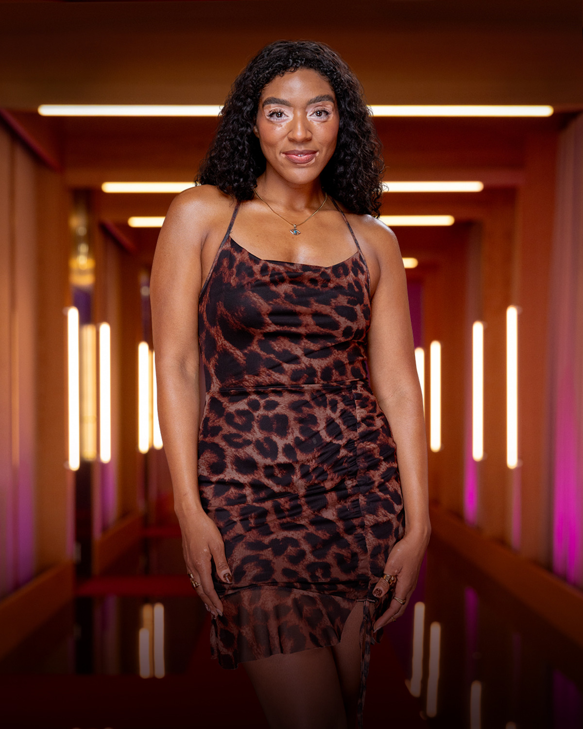 Woman with curly hair wearing a leopard print dress stands confidently in a modern hallway with warm lighting and vertical light fixtures, creating a stylish, contemporary atmosphere.